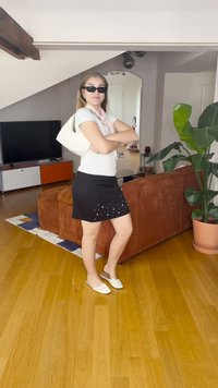 Bela torbica, kratka črna krila z bisernimi poudarki, bela majica in ravne čevlje. Ozadje vsebuje leseno tla, rjavo sof in rastlino.