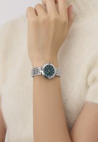 Ezüst óra kerek zöld számlappal, kristálydíszekkel és texturált fém karkötővel, egy kézen viselve, világos pulóver háttér előtt.