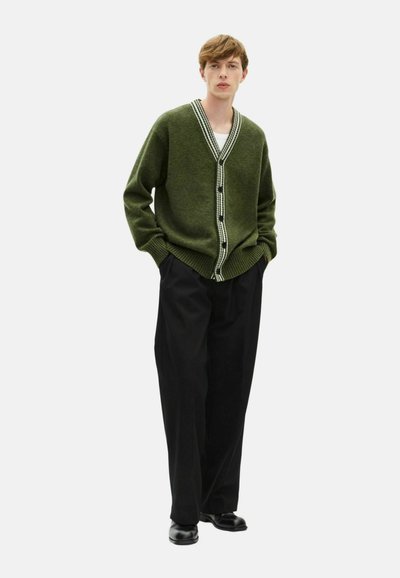 Cardigan verde con scollatura a V, rifiniture a strisce bianche a contrasto e bottoni neri, abbinato a pantaloni neri a gamba larga e scarpe nere.