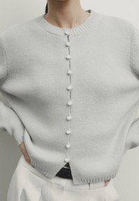 Harmaa neulepusero, jossa pyöreä pääntie, edessä nappikiinnitys tupsukoristein ja löysä istuvuus. Rapeakuvioiset resorihihaiset.