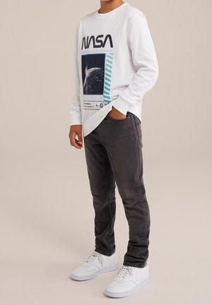 Garçon portant un t-shirt blanc à manches longues NASA avec un graphisme de la Terre, un jean gris foncé et des baskets blanches, debout avec une main dans la poche.