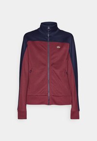 Veste zippée avec col haut, corps bordeaux, manches et épaules bleu marine, poches latérales zippées et petit logo crocodile sur la poitrine.