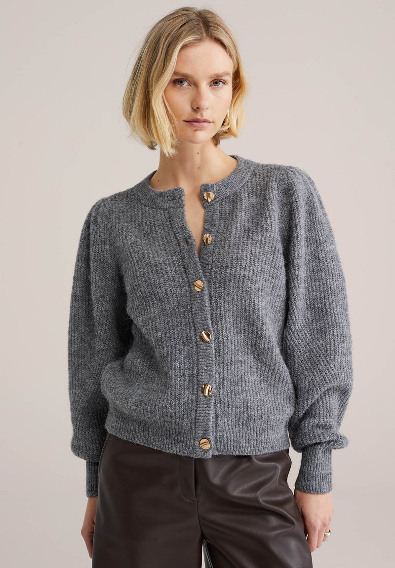 Grå stickad kofta med rund halsringning, ribbad struktur och sex knappar i guldfärg framtill. Puffärmar och åtsittande muddar.