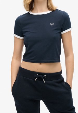 Kobieta w granatowym, krótkim T-shircie z białym wykończeniem i logo "Super Dry", w granatowych spodniach dresowych ze ściągaczem, ręce w kieszeniach.