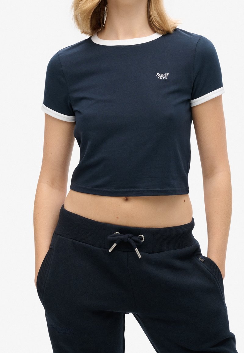 Femme portant un t-shirt court bleu marine avec bordure blanche et logo "Super Dry", associé à un pantalon de jogging bleu marine avec cordon de serrage, mains dans les poches.