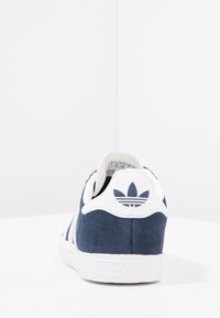 Mørkeblå semsket sneakers med hvite detaljer, med tre hvite striper og en teksturert gummisåle. Logoet på hælen er synlig.