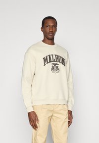 Κρεμ σμπώτο με στρογγυλή λαιμόκοψη και μακριά μανίκια, με εκτυπωμένο το "MALBON" σε σκούρο χρώμα και ένα γραφικό έμβλημα από κάτω.