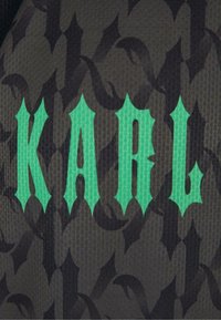 Juodas tekstūruotas audinys su tamsiu raštu, kuriame išryškėja ryškus, stilizuotas žalias tekstas, rašantis "KARL" kampuotu šriftu.