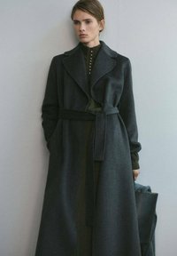 Lungo cappotto in lana grigio scuro con cintura, ampio colletto e rever, indossato sopra una maglia verde scuro con dettagli a bottoni.