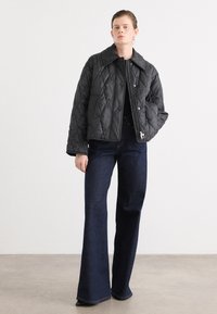 Zwarte gewatteerde jas met een brede kraag, voorzien van twee knopen en een decoratieve accent. Gepaard met donkerblauwe flared jeans.