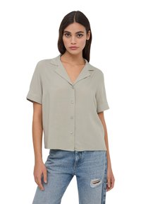 Blusa grigia chiara a maniche corte con colletto, chiusura a bottoni e vestibilità comoda, realizzata in un tessuto liscio. Indossata con jeans blu strappati.