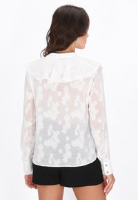 Blusa blanca translúcida con patrón floral texturizado, cuello con volantes y mangas largas con puños abotonados. Combinada con pantalones cortos negros.