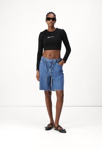 Čierny dlhý rukáv cropped top s logom, spárovaný so svetlo modrými džínovými kraťasmi so sťahovacou šnúrkou a rozstrapkaným lemom. Nosené s čiernymi sandálmi.
