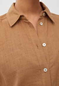 Chemise boutonnée couleur beige avec un tissu texturé, dotée d'un col et de deux boutons ronds. Le tissu semble léger et respirant.