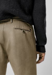 Beige broek van gladde stof, met een achterzak met knoopsluiting, gecombineerd met een donkere gebreide trui met structuur.
