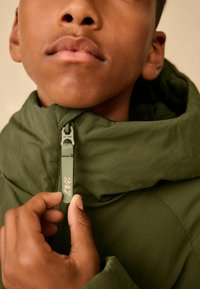 Grüne Zip-Jacke mit hohem Kragen und strukturiertem Stoff. Verfügt über einen sichtbaren Reißverschluss mit der Nummer "247." Nahaufnahme einer Hand, die den Reißverschluss hält.