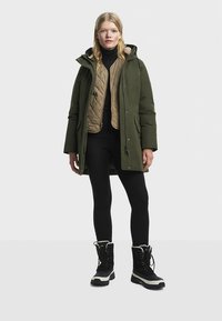 Padded green parka with a hood, tan quilted vest underneath, black turtleneck, black leggings, and black winter boots with white soles.