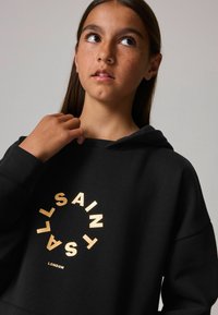 Sudadera con capucha negra con un estampado circular en foil dorado que dice "SAINT LONDON". Presenta puños acanalados y un corte relajado.