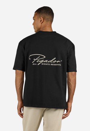 Man facing away wearing a black T-shirt with "Pegador All Rights Reserved" text on the back, paired with light-colored pants.