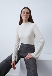 Langärmliges, cremefarbenes, geripptes Oberteil mit hohem Kragen, kombiniert mit hochtaillierten, strukturierten grauen Hosen; die Hand des Models ruht auf dem Oberschenkel.