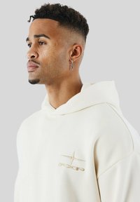 Giovane con capelli corti e ricci e piercing sul viso, che indossa un felpa leggera beige con un piccolo logo ricamato sul petto.