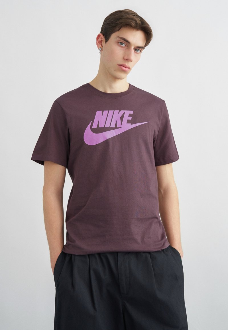 Jonge man die een paarse Nike T-shirt met roze logo draagt, zwarte broek, staand tegen een effen lichtgrijze achtergrond.