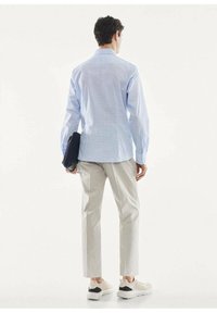Homme portant une chemise bleu clair, un pantalon beige et des baskets blanches, tenant un sac d'ordinateur portable noir, debout de dos sur un fond blanc.