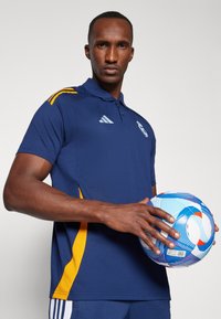 Námořnická modrá polo tričko s žlutými akcenty, které má límec a tři knoflíky. Model drží modro-oranžový fotbal.