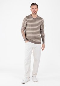 Hombre con brazos tatuados que lleva una camisa de mangas cortas de rayas marrón y beige, pantalones blancos y zapatillas blancas, de pie contra un fondo blanco.