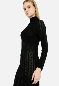 Robe en tricot noire à manches longues, avec un col haut, un motif texturé et des découpes sur les côtés.