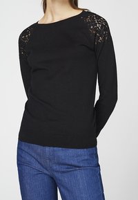 Musta neulepusero, jossa pitsikoristeet olkapäillä, resorikahvat ja -helma. Pitkät hihat ja pyöreä pääntie. Yhdistettysinin farkkujen kanssa.