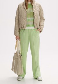 Beige vadderad jacka, mönstrad grön tröja, ljusgröna byxor och beige dragkedjeväska med varumärke. Vita sneakers med gröna accenter.