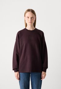 Sudadera de color morado oscuro con cuello redondo y mangas largas, que presenta un sutil texto bordado en la parte frontal. Combinada con jeans de color azul claro.