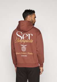 Brązowa bluza z kapturem z dużą białą i żółtą grafiką na plecach, z tekstem i emblematem. Miękki materiał, luźny krój i kaptur ze sznurkiem.