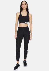 Kobieta w czarnym staniku sportowym i legginsach z logo FILA, stojąca z neutralnym wyrazem twarzy na jednolitym tle.