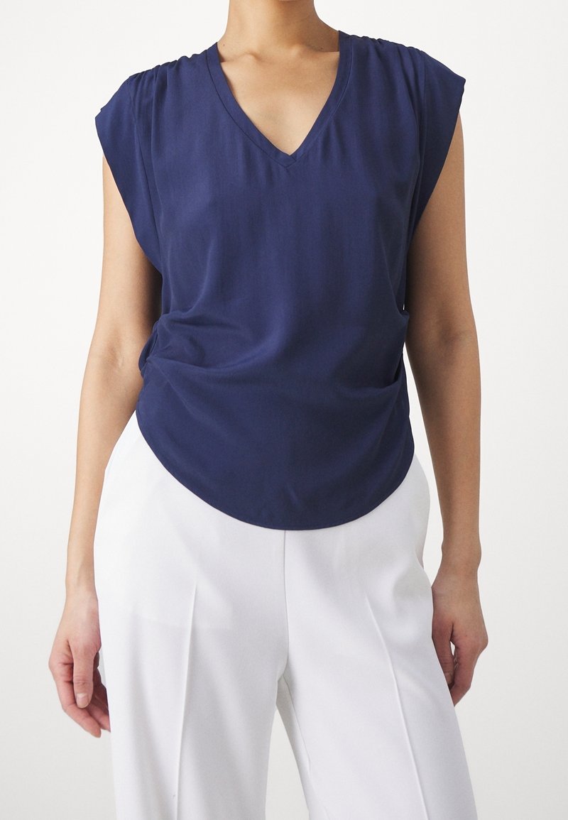 Femme portant un haut bleu marine sans manches à col en V associé à un pantalon blanc taille haute à jambes larges sur un fond uni.