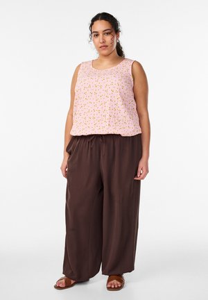 Moteris su garbanotais plaukais, vilkinti rožinę be rankovių palaidinę su mažais geltonais žiedais, laisvas rusvas kelnes ir rudas sandalus, stovinti prieš baltą foną.