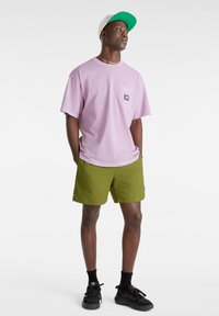 T-shirt viola chiaro con taschino e logo, abbinato a pantaloni corte verde oliva, calzini neri e sneaker nere. Sfondo bianco.