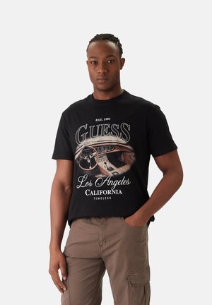 Hombre con el cabello trenzado que lleva una camiseta negra gráfica con el interior de un coche vintage y el texto "Guess Los Ángeles California Atemporal", pantalones marrones.