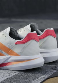 Chaussures de course blanches avec des accents orange et rose reposant sur une surface texturée, affichant les talons et une partie des côtés.