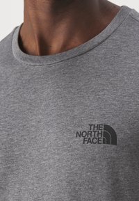 Detailní záběr osoby v šedém tričku s černým logem The North Face na hrudi, ukazující dekolt a oblast ramen.