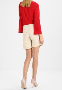 Haut rouge à manches longues associé à un short beige orné de broderies d'étoiles. Le short arrive au genou et la tenue est complétée par des talons nus.