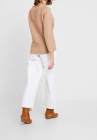 Beige ribstrikket sweater med lange ærmer, figursyet ved taljen, kombineret med hvide bukser med vide ben og lysebrune ankelstøvler. Glat tekstur, minimalistisk design.