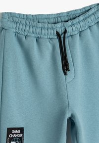 Lichtblauwe joggers met een trekkoord in de taille, voorzien van een zijpatch met de tekst "GAME CHANGER." Soepele, katoenmix stof.