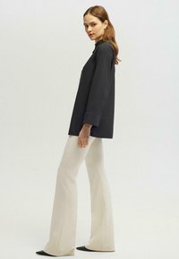 Camisa negra de gran tamaño con cuello alto, mangas largas y puños abotonados, combinada con pantalones acampanados de color blanco roto y zapatos negros de punta.