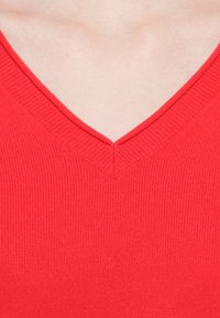 Suéter rojo de cuello en V con textura acanalada, que muestra un acabado suave y un borde de cuello definido y ordenado. Primer plano de la tela y el escote.