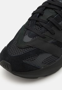 Black athletic shoe featuring mesh and suede materials, layered design with textured accents, laces, and a cushioned sole.