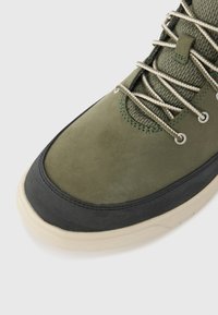 Olive green athletic shoe with a suede upper, black rubber toe cap, and mesh accents. Features cream-colored laces and sturdy eyelets.