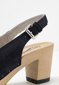 Marinblå mocka klacksandaler med en träklack och justerbart ankelsnöre med silverfärgad hårdvara och kontrasterande sömdetaljer.