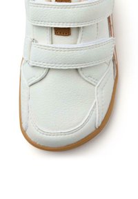 Zapato de piel sintética blanca con dos correas de velcro, punta redondeada y suela de goma marrón. Detalles de costura simples en la parte superior.